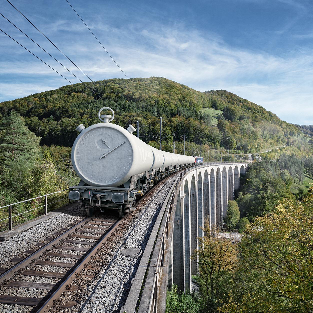 Vigier Ciment Keyvisual: Ein Zug ist mit einer riesigen Beton-Stoppuhr beladen und fährt über eine Brücke.
